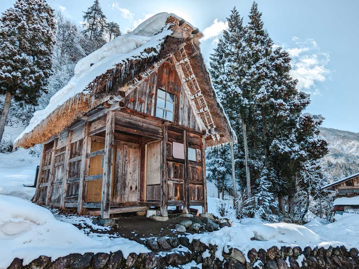 Small Gassho house covered in snow and icicles in front of snowy forest in Shirakawago.