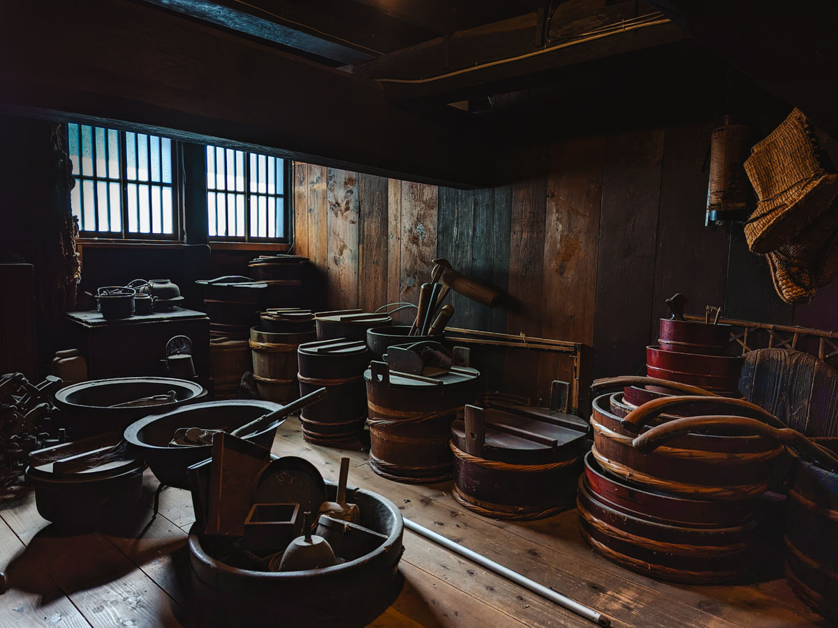 Dark interior of Wada House room with old wooden barrels and various cooking tools.