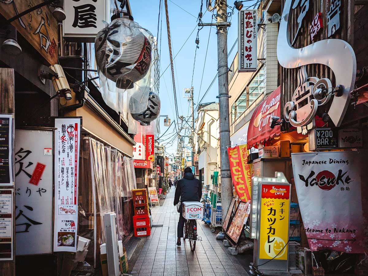 Man cycling down narrow Tokyo alley lined with store signs and lanterns.