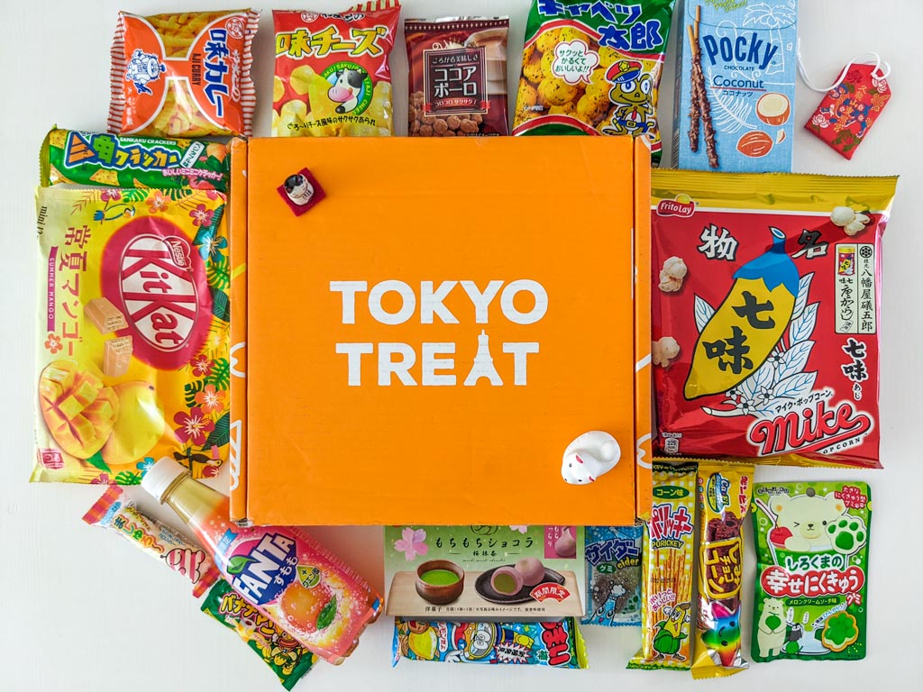 Orange TokyoTreat box surrounded by Japanese snacks on white table.