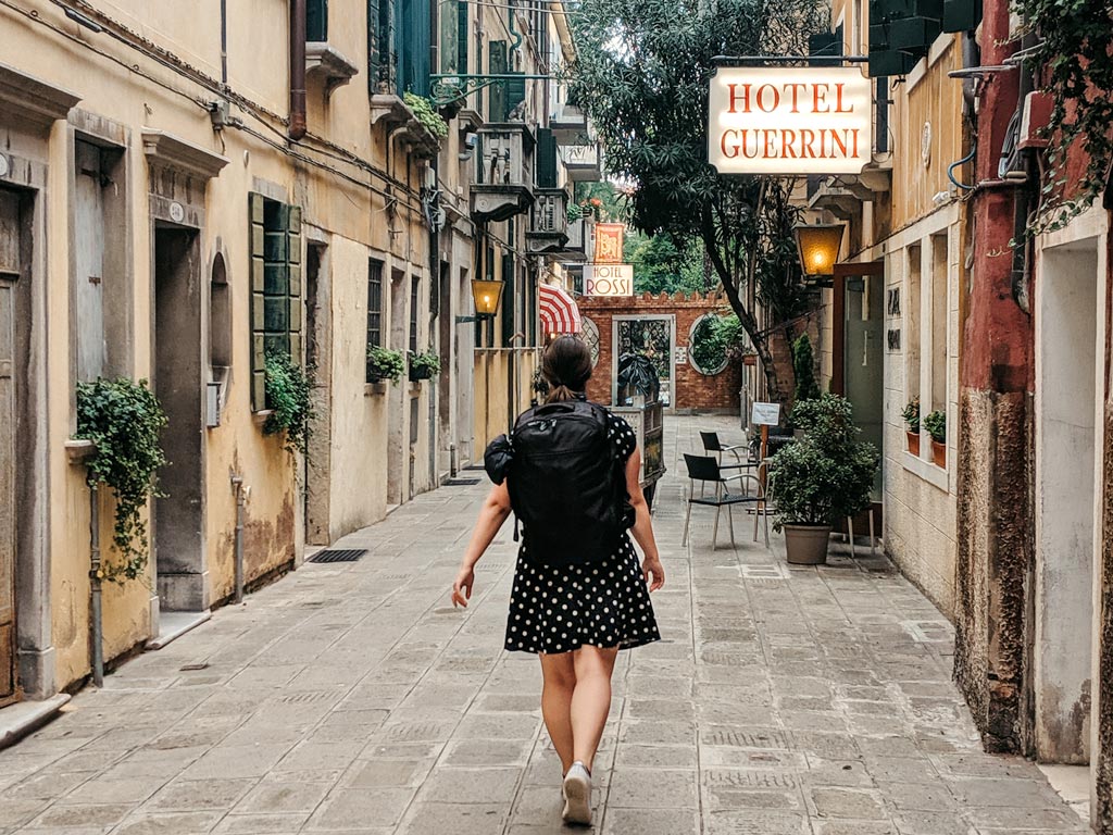 Woman walking down street in Venice wearing black travel backpack and dress.