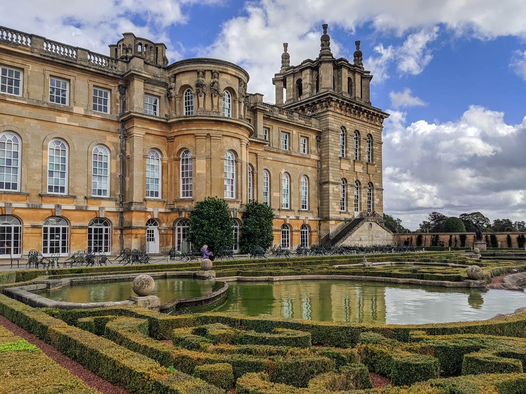 Hedge gardens and pond in front of Blenheim Palace stone exterior.