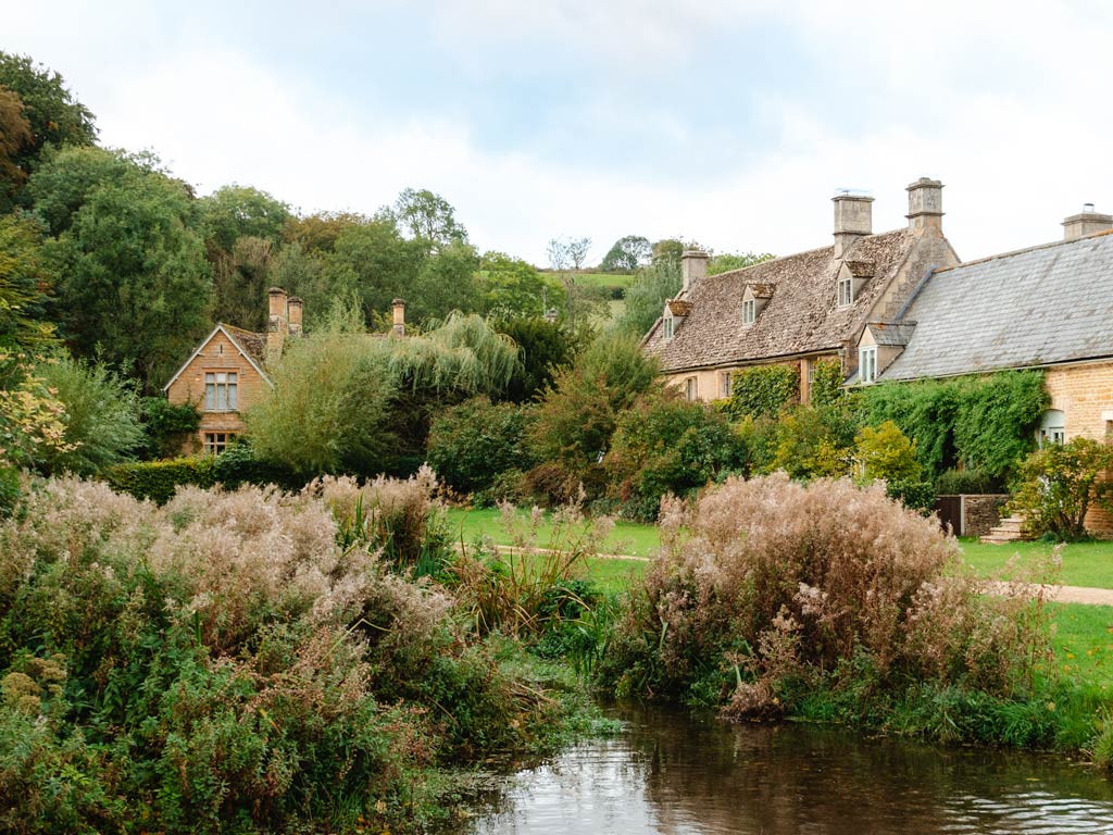 Cotswolds stone cottages with pond and foliage in foreground.