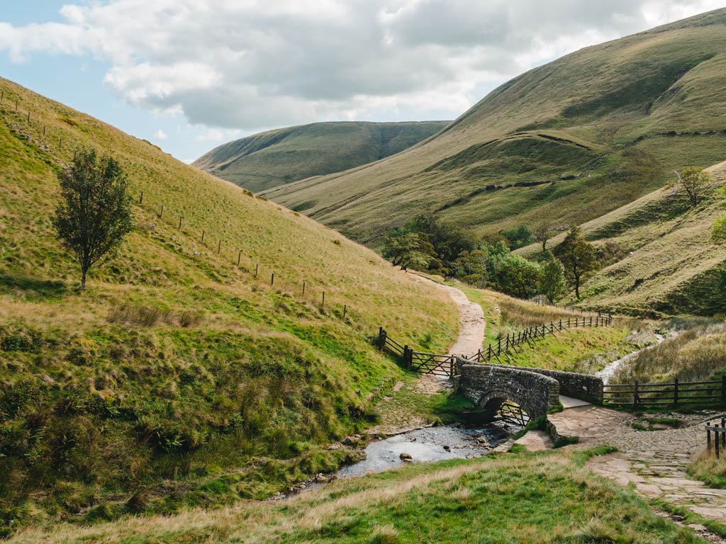 Rolling green hills of UK Peak District with stone bridge crossing small stream.