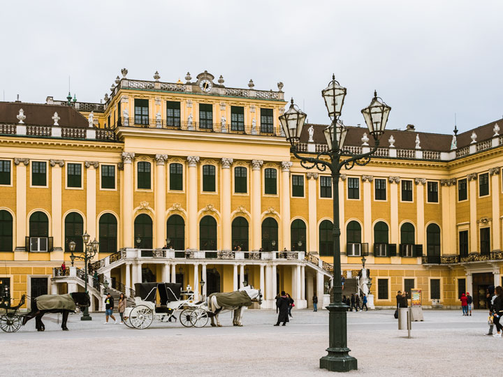 Yellow facade of Schonbrunn Palace with horse carriages and lamp post.